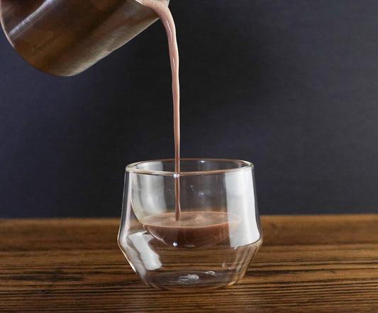 Jug pouring a 100% organic hot chocolate into a clear glass on a wooden surface with a dark background