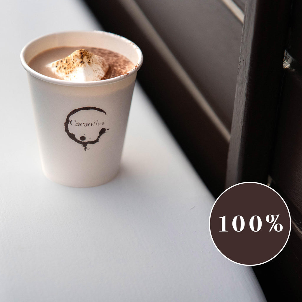 Close up of a cacao brew branded paper cup containing luxury organic hot chocolate and a mega mallow. Cup is placed on the mudguard of the Cacao Brew drinks trailer. A circular sign shows 100% which indicates the percentage of cacao powder in this drink.