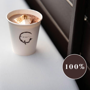 Close up of a cacao brew branded paper cup containing luxury organic hot chocolate and a mega mallow. Cup is placed on the mudguard of the Cacao Brew drinks trailer. A circular sign shows 100% which indicates the percentage of cacao powder in this drink.