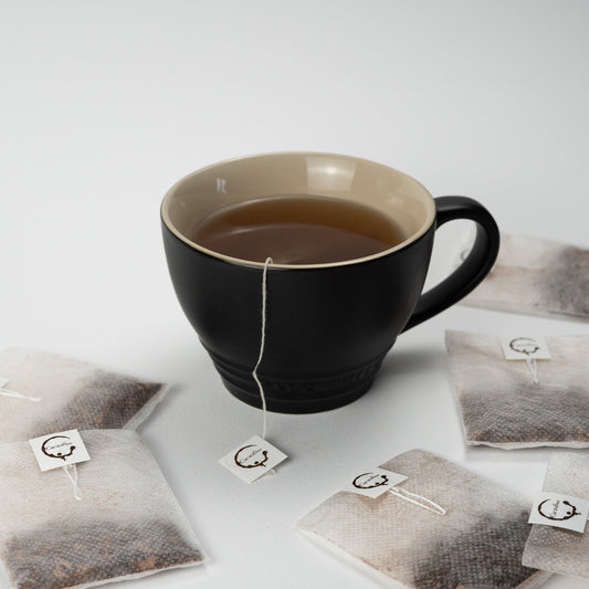 A cup of cacao chocolate tea with several biodegradable brew bags scattered around on a white surface.