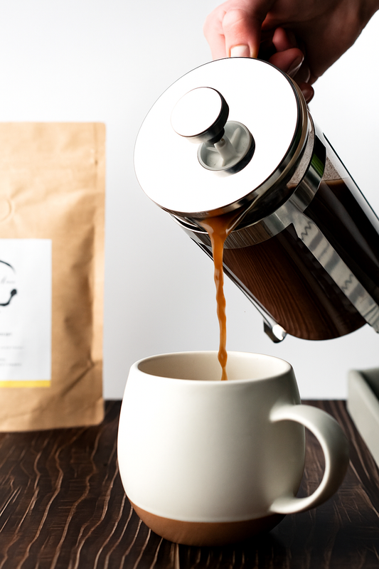 Person pouring Philippine brewed cacao from a French press into a white mug with a 1kg bag of Cacao Brewed grounds in the background.